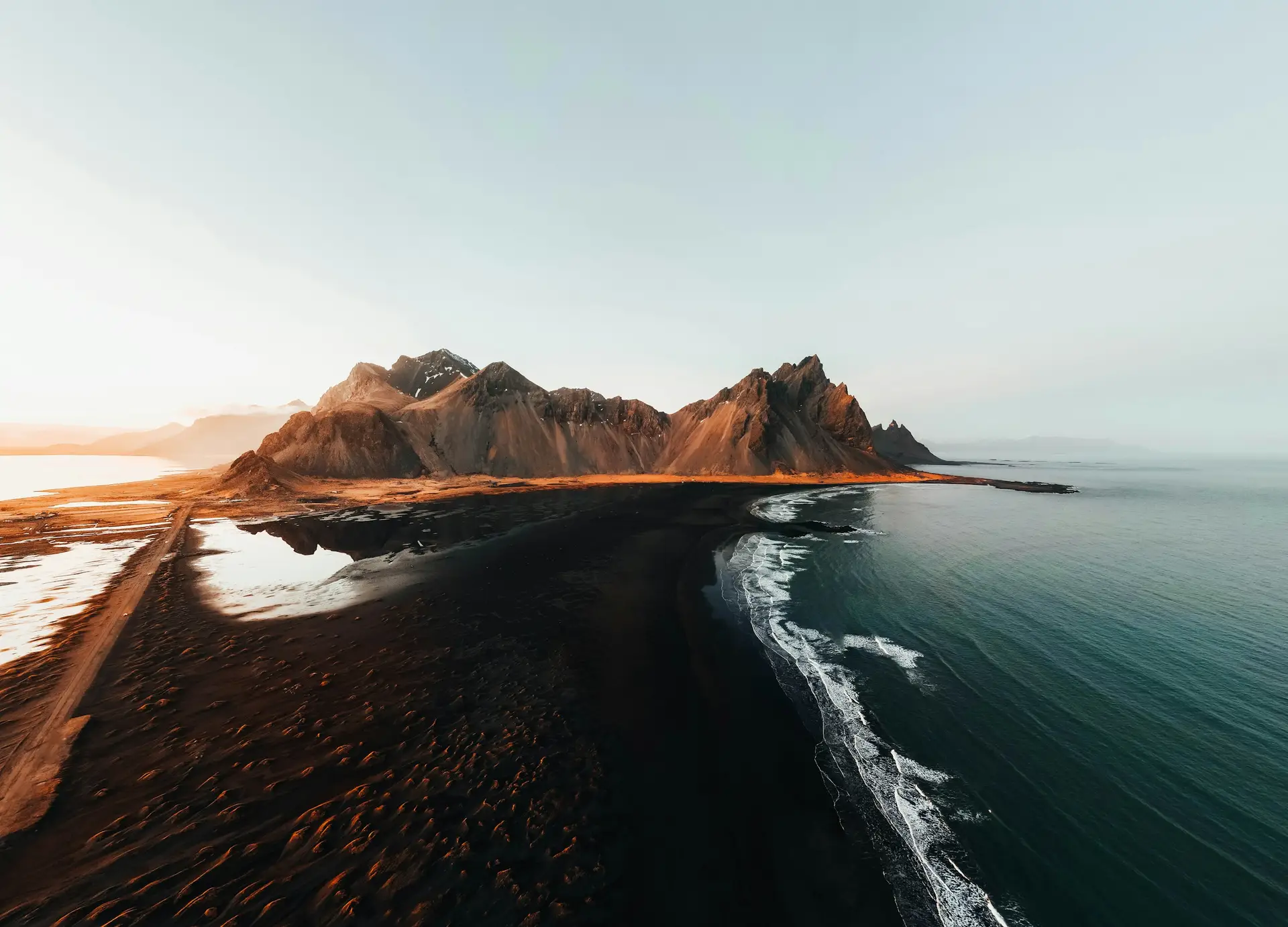 Luftaufnahme eines Bergmassivs ueber schwarzem Sandstrand im Abendlicht