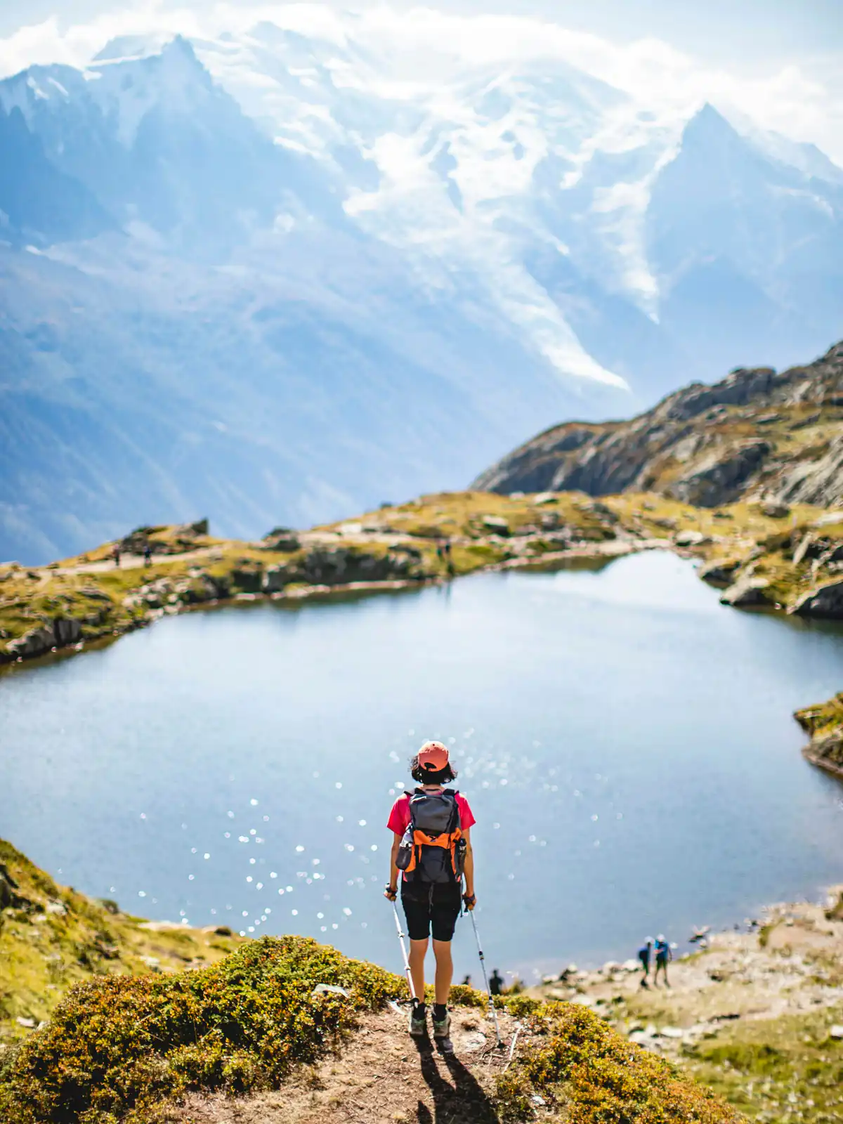 Ein Mensch mit Rucksack und Wanderstöcken steht klein über einem Bergsee, dahinter verschneite Gipfel
